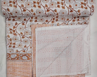 Puede incluir: Una colcha de algodón blanco con un patrón floral repetitivo de contornos marrones y azules. La tela presenta un borde de rayas horizontales marrones y un reverso de tela blanca con detalles visibles de bordado en punto hilván naranja.