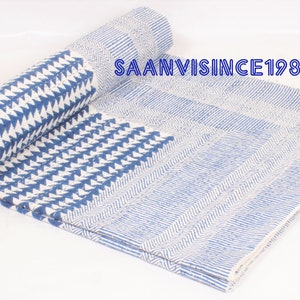 May include: A folded textile featuring a blue and white geometric pattern with repeating triangles and striped sections. The text SAANVISINCE1988 is printed in blue on the fabric. The material displays a quilted texture with visible stitching.