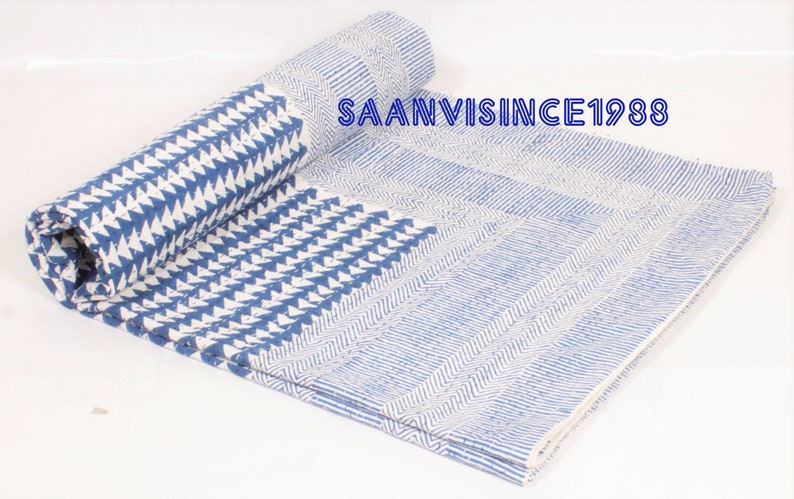 May include: A folded textile featuring a blue and white geometric pattern with repeating triangles and striped sections. The text SAANVISINCE1988 is printed in blue on the fabric. The material displays a quilted texture with visible stitching.