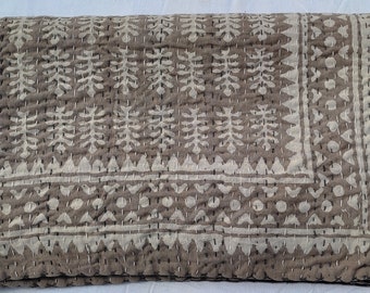 May include: A folded, brown and white patterned quilt. The quilt features a repeating leaf and geometric design, with white stitching throughout. The edges are bordered with a decorative pattern.