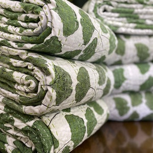 May include: A stack of folded white quilts featuring a repeating green ginkgo leaf pattern. The fabric is detailed with visible horizontal and vertical running stitch quilting lines in a matching green thread throughout the entire surface.