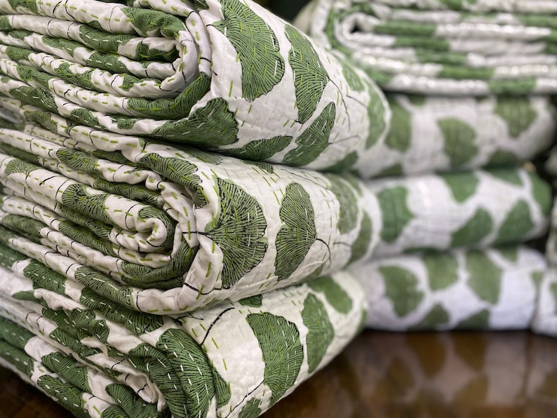 May include: A stack of folded white quilts featuring a repeating green ginkgo leaf pattern. The fabric is detailed with visible horizontal and vertical running stitch quilting lines in a matching green thread throughout the entire surface.