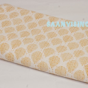 May include: White fabric with a yellow leaf pattern. The fabric is folded in half and the text "SAANVISINCE1988" is printed in blue on the top right corner.