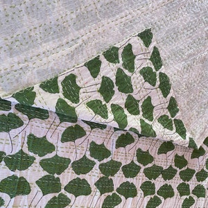 May include: A white textile featuring a repeating pattern of dark green ginkgo leaves. The fabric is layered and folded, revealing a textured surface with visible horizontal running stitches in a light green thread throughout the material.