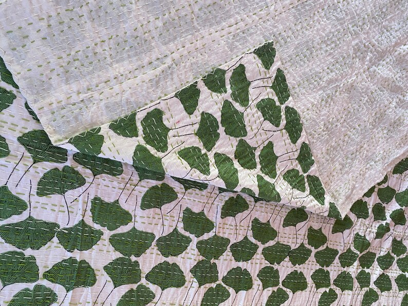 May include: A white textile featuring a repeating pattern of dark green ginkgo leaves. The fabric is layered and folded, revealing a textured surface with visible horizontal running stitches in a light green thread throughout the material.