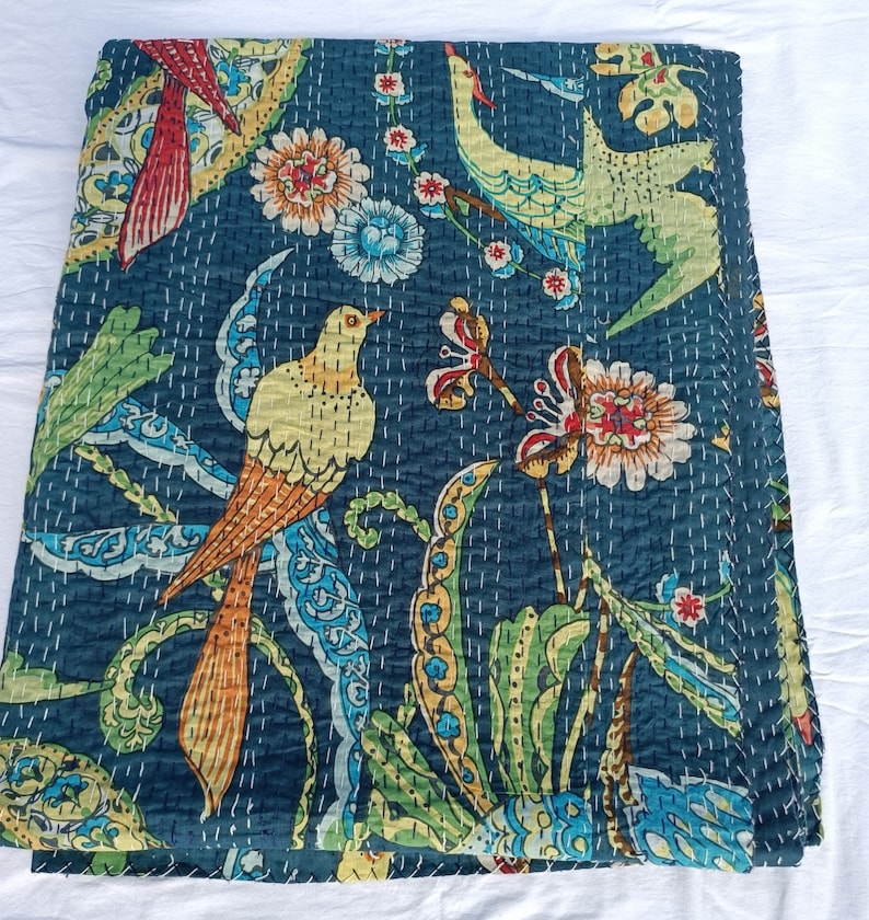 May include: A folded, dark teal quilt with a vibrant bird and floral pattern. The design features colorful birds with yellow, orange, and teal feathers, surrounded by flowers and foliage in shades of green, blue, and red. White stitching outlines the design.