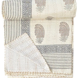 May include: A folded off-white cotton quilt featuring a repeating pattern of brown paisley motifs, dark gray geometric borders, and vertical stripes. The textile is finished with visible running stitch embroidery in a light tan thread throughout.
