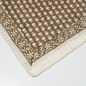May include: A brown textile featuring a repeating white floral block print pattern. The fabric is finished with a wide white border decorated with a white floral vine motif and is held together by visible straight running stitch quilting lines.