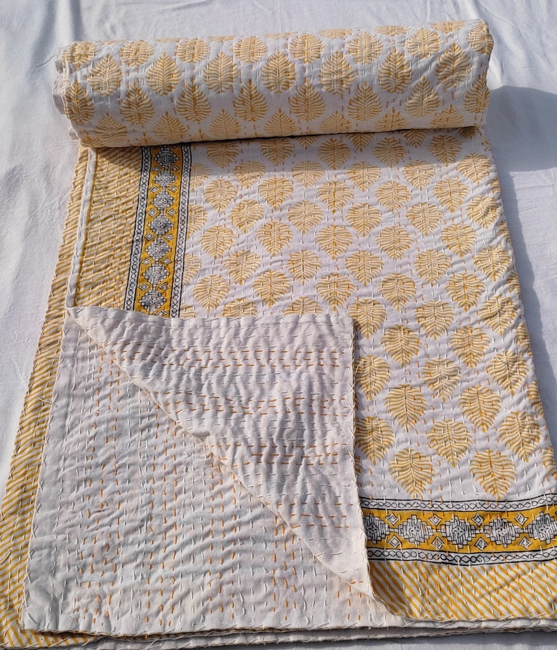 May include: A white and yellow quilt with a leaf pattern. The quilt is folded and rolled, showcasing the intricate stitching and design. The border features a geometric pattern with yellow and black accents. The quilt is made of soft, comfortable material.