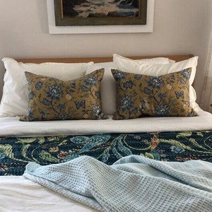 May include: Two decorative throw pillows with a mustard yellow background and blue floral patterns rest on a bed. The bed is made with white sheets, a patterned runner, and a light blue waffle-weave blanket.