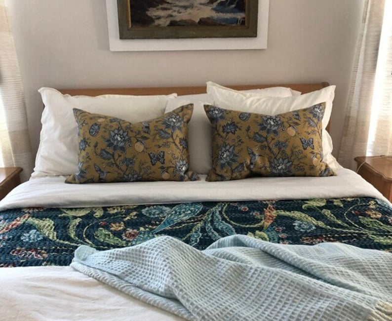 May include: Two decorative throw pillows with a mustard yellow background and blue floral patterns rest on a bed. The bed is made with white sheets, a patterned runner, and a light blue waffle-weave blanket.