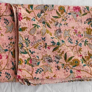 Op de afbeelding: Een gevouwen quilt met een bloemenpatroon in roze, groen en wit. Het ontwerp heeft een herhalend patroon van bloemen en bladeren, met zichtbare stiksels. De zachte kleuren en het ontwerp van de quilt zouden een vleugje natuur toevoegen aan elke kamer.