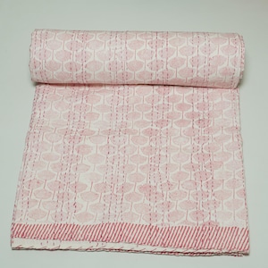 May include: A folded quilt featuring a white base with a repeating pink ginkgo leaf pattern. The textile displays vertical rows of running stitches in a matching pink thread, with a striped border along the bottom edge of the fabric.