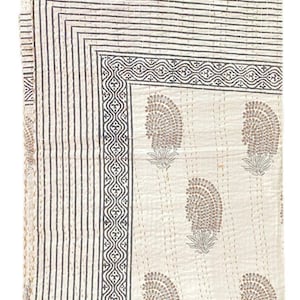 May include: A rectangular off-white cotton quilt featuring a repeating brown paisley-style floral pattern. The border consists of multiple thin dark brown parallel lines and a decorative geometric band. The fabric has visible running stitch details.