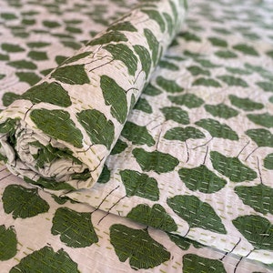 May include: A white quilt featuring a repeating pattern of green ginkgo leaves. The fabric is decorated with visible, straight-line running stitches in a light green thread that follow the contours of the leaves and the overall textile surface.