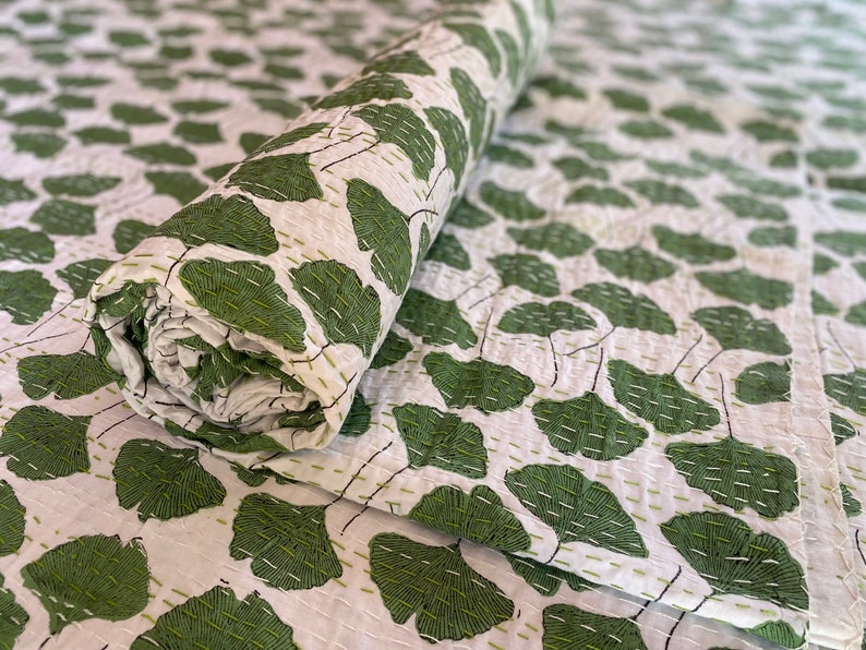 May include: A white quilt featuring a repeating pattern of green ginkgo leaves. The fabric is decorated with visible, straight-line running stitches in a light green thread that follow the contours of the leaves and the overall textile surface.