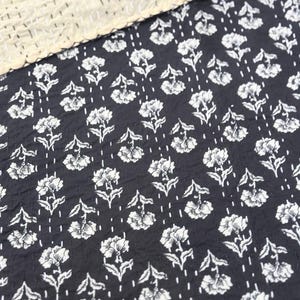May include: A black and white floral patterned textile. The design features white flowers with stems and leaves, arranged in rows. The top edge has a cream-colored, textured border with white stitching. The fabric appears to be cotton or a similar material.