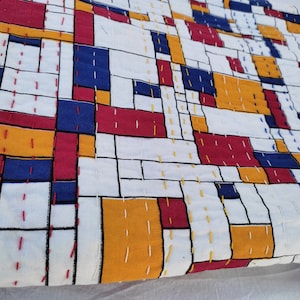 May include: A white fabric with a geometric pattern of red, blue, and yellow squares. The fabric is quilted with white thread.