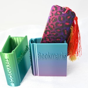 May include: Three colorful bookmark holders with the word "Bookmarks" printed on them. The holders are made of plastic and have a gradient color scheme. One holder is green, one is blue, and one is purple. A leopard print bookmark is placed in the blue holder.