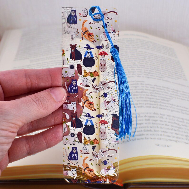 May include: A clear bookmark with a blue tassel. The bookmark features a repeating pattern of cats dressed in Halloween costumes. The cats are wearing witch hats, vampire capes, and other costumes. The bookmark also features pumpkins, bats, and other Halloween-themed imagery.
