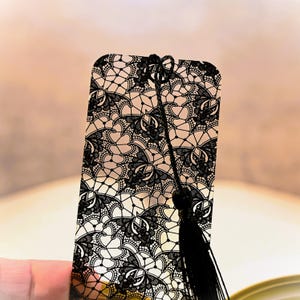 May include: A black lace bookmark with a tassel. The bookmark is made of a clear material with a black lace design.