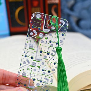 Puede incluir: Marcapáginas transparente con un diseño temático de golf. El patrón incluye pelotas de golf, carritos de golf y palos en verde y azul. Una borla verde cuelga de la parte superior. El marcapáginas está hecho de plástico transparente.