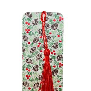 May include: A rectangular bookmark with a festive pattern of pinecones, green pine needles, and red berries. A long, red tassel is attached to the top. The background is white.