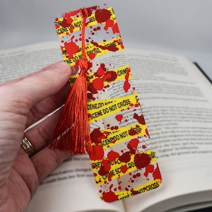 May include: A red and yellow bookmark with a red tassel. The bookmark has a crime scene do not cross tape design with red blood splatter.