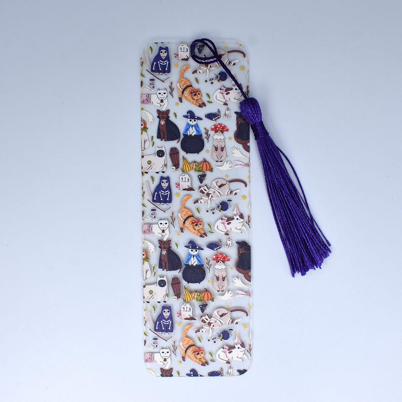 May include: A clear acrylic bookmark with a purple tassel. The bookmark features a repeating pattern of cats dressed in Halloween costumes, including witches, skeletons, and pumpkins.