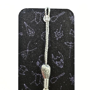 May include: A black bookmark with a silver tassel and a constellation pattern. The constellations are printed in white and are scattered across the bookmark.