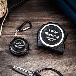 May include: Two black measuring tapes with a silver face and white text. The larger tape reads "Tool of Measuring +4 perception wt: 7.2". The smaller tape reads "Tool of Measuring +4 perception wt: 1.5".