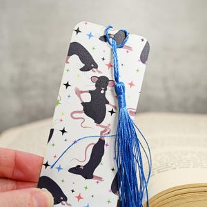 May include: A white bookmark with a blue tassel. The bookmark features a repeating pattern of black rats in various yoga poses on a white background with colorful stars.