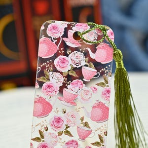 May include: A decorative bookmark featuring a floral pattern of pink roses, teacups, and teapots on a white background. The bookmark has a green tassel attached to the top. The design is repeated across the bookmark.