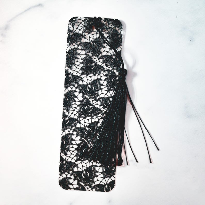 May include: Black and white lace bookmark with a black tassel. The bookmark is made of metal and has a delicate lace design.