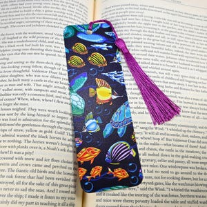 May include: Bookmark featuring a vibrant underwater scene with colorful fish, a sea turtle, and bubbles against a dark blue background. It has a purple tassel. The bookmark is designed to be inserted into a book to mark a reader's place.