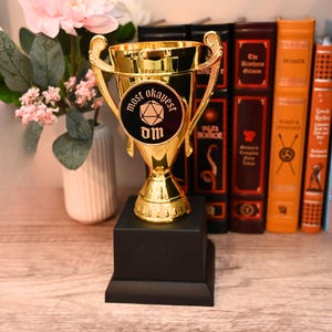 May include: A gold-colored trophy with the words "most okayest" and "dm" on a black circular emblem. The trophy sits on a black base, with a floral arrangement and a row of books in the background.