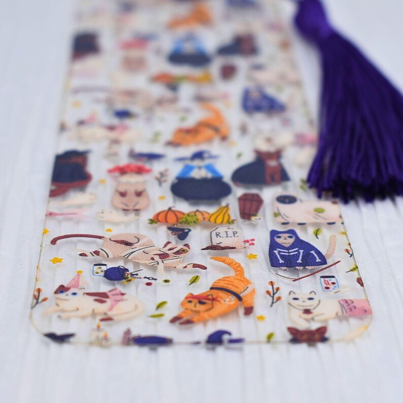 May include: A clear acrylic bookmark with a Halloween cat theme. The bookmark features a variety of cats dressed in costumes, including a witch, a skeleton, and a vampire. The bookmark also features pumpkins, bats, and other Halloween-themed imagery. The bookmark has a purple tassel.