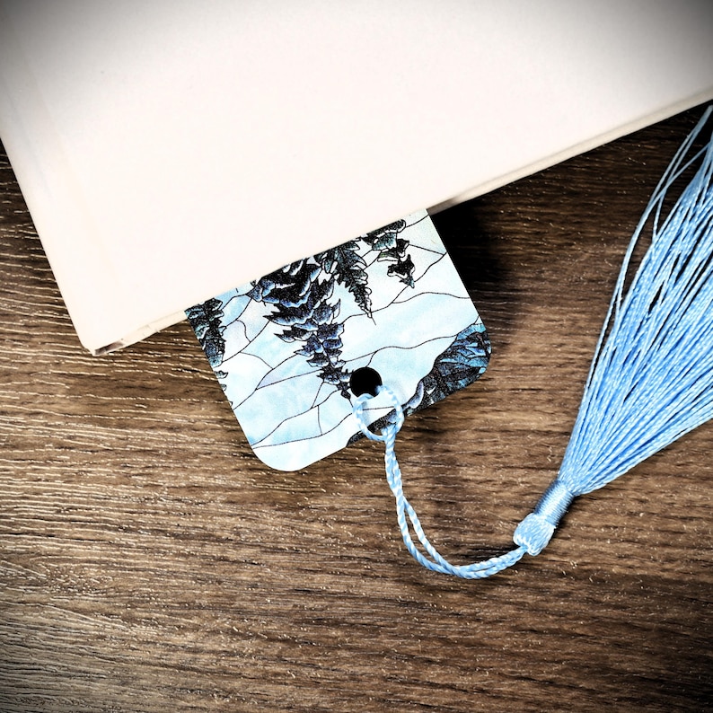 May include: A blue and white bookmark with a floral design and a blue tassel. The bookmark is made of paper and has a hole punched in the top for a string.