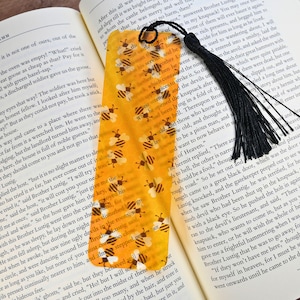 May include: A yellow bookmark with a bee pattern and a black tassel. The bookmark is rectangular with a rounded top and is placed on an open book.
