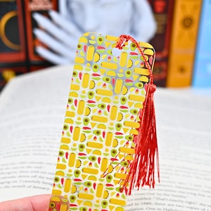 Puede incluir: Un marcapáginas dorado con un borla roja. El marcapáginas presenta un patrón repetido de tacos amarillos, aguacates verdes y chiles rojos.
