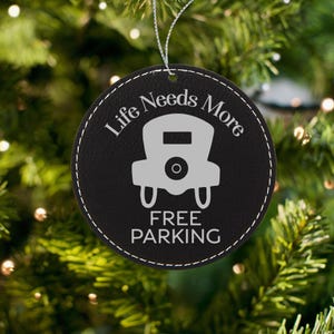 Może przedstawiać: Czarna skórzana ozdoba choinkowa z białymi szwami. Ozdoba jest okrągła i przedstawia grafikę zabytkowego samochodu z tekstem "Life Needs More Free Parking".