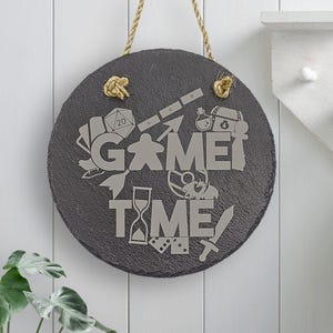 Peut inclure: Une pancarte ronde en ardoise noire avec un cintre en corde. La pancarte indique "Game Time" en lettres blanches et est d&eacute;cor&eacute;e de diverses ic&ocirc;nes li&eacute;es aux jeux, notamment un d&eacute; &agrave; 20 faces, une pi&egrave;ce de jeu, une &eacute;p&eacute;e et un sablier.
