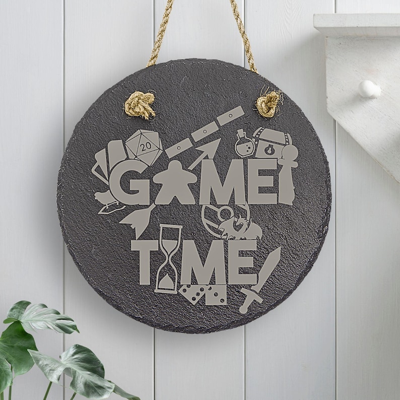 Peut inclure: Une pancarte ronde en ardoise noire avec un cintre en corde. La pancarte indique "Game Time" en lettres blanches et est d&eacute;cor&eacute;e de diverses ic&ocirc;nes li&eacute;es aux jeux, notamment un d&eacute; &agrave; 20 faces, une pi&egrave;ce de jeu, une &eacute;p&eacute;e et un sablier.