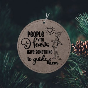 May include: A round, brown leather ornament with a stitched edge. It features a black and white illustration of the Tin Man from the Wizard of Oz, along with the text "People with Hearts have something to guide them".