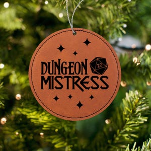 May include: A brown leather Christmas ornament with the text "Dungeon Mistress" and a black 20-sided die graphic.