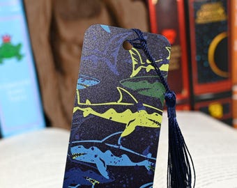 May include: A rectangular bookmark with a navy blue background and a pattern of sharks in shades of blue, green, and yellow. A navy blue tassel hangs from the top. The bookmark is placed in an open book.