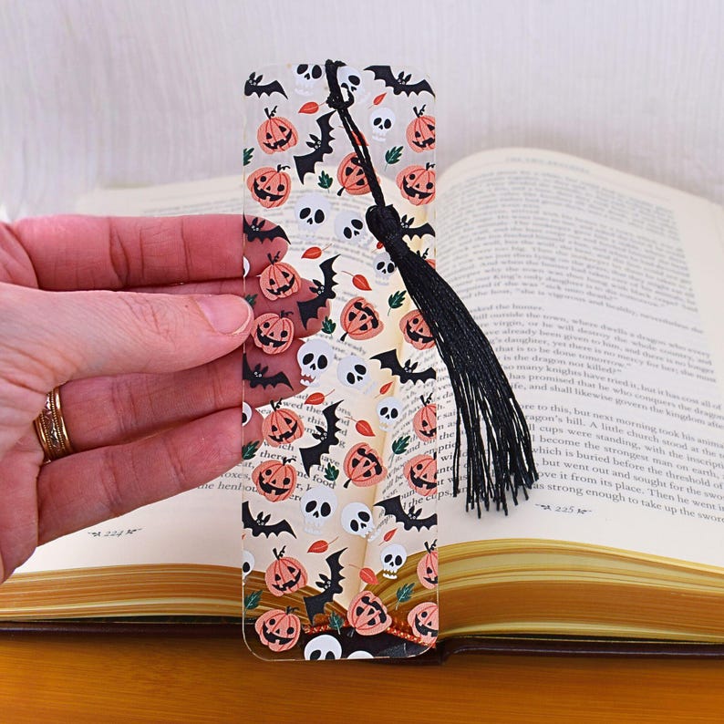May include: A clear glass bookmark with a black tassel. The bookmark features a repeating pattern of orange pumpkins, black bats, and white skulls on a white background.