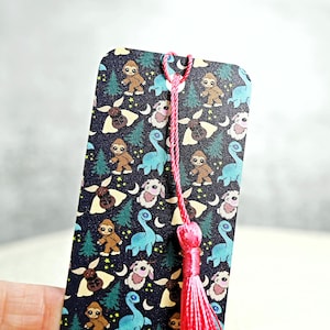 May include: A black bookmark with a pink tassel. The bookmark features a repeating pattern of blue dinosaurs, brown sasquatch, and white moths on a dark background.