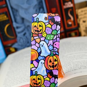 Puede incluir: Un marcapáginas con temática de Halloween con calabazas, fantasmas y una bruja. El diseño de estilo vidriera utiliza colores naranja, amarillo, morado y azul. Tiene una borla naranja y está sobre libros abiertos.