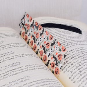 May include: A clear plastic bookmark with a Halloween theme. The bookmark features a repeating pattern of orange pumpkins, black bats, and white skulls. The bookmark has a black tassel.